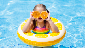 Young girl in a yellow ring in the swimming pool holding two half oranges to her eyes.
