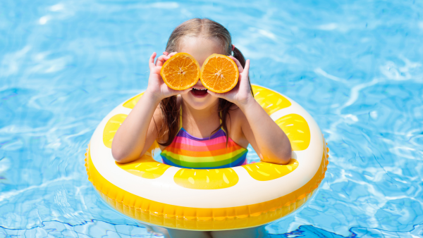 Young girl in a yellow ring in the swimming pool holding two half oranges to her eyes.